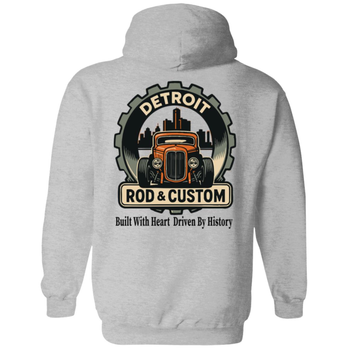 Back of a gray hoodie with a large round gear badge: "DETROIT" arched at top, an orange chopped hot rod coupe in front of a black Detroit skyline, a "ROD & CUSTOM" banner, and "Built With Heart Driven By History" in script below.