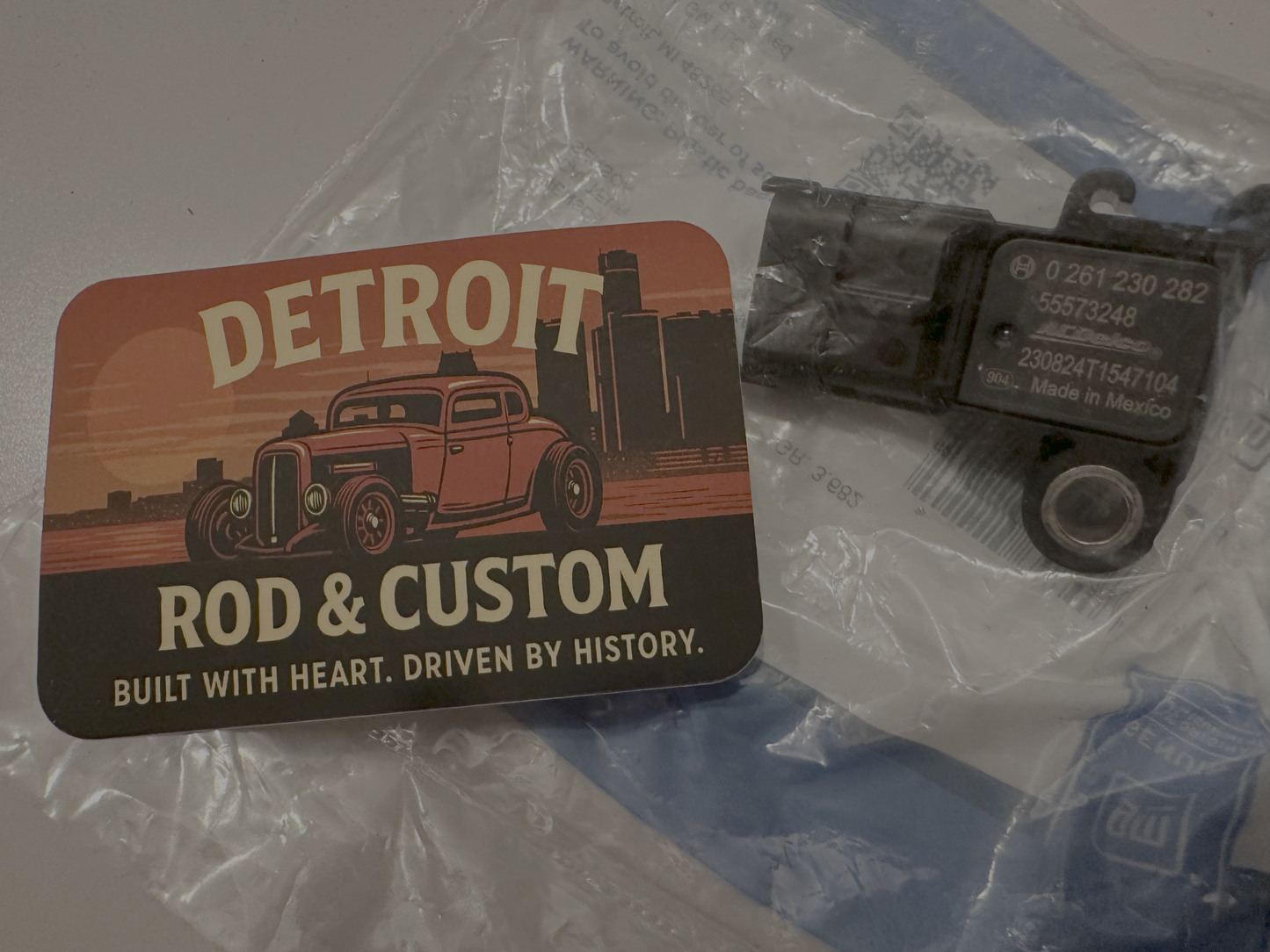 Small mechanical component on a clear plastic sheet with 'Rod & Custom Detroit' branding.