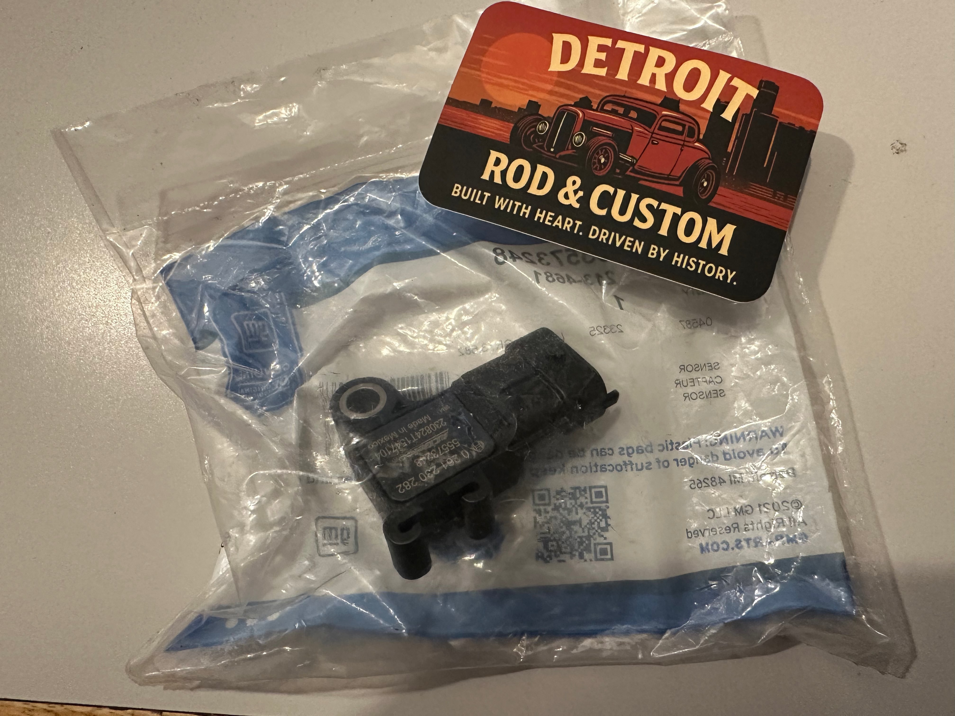 Black mechanical part in clear plastic packaging with a 'Detroit Rod & Custom' card on a gray surface.