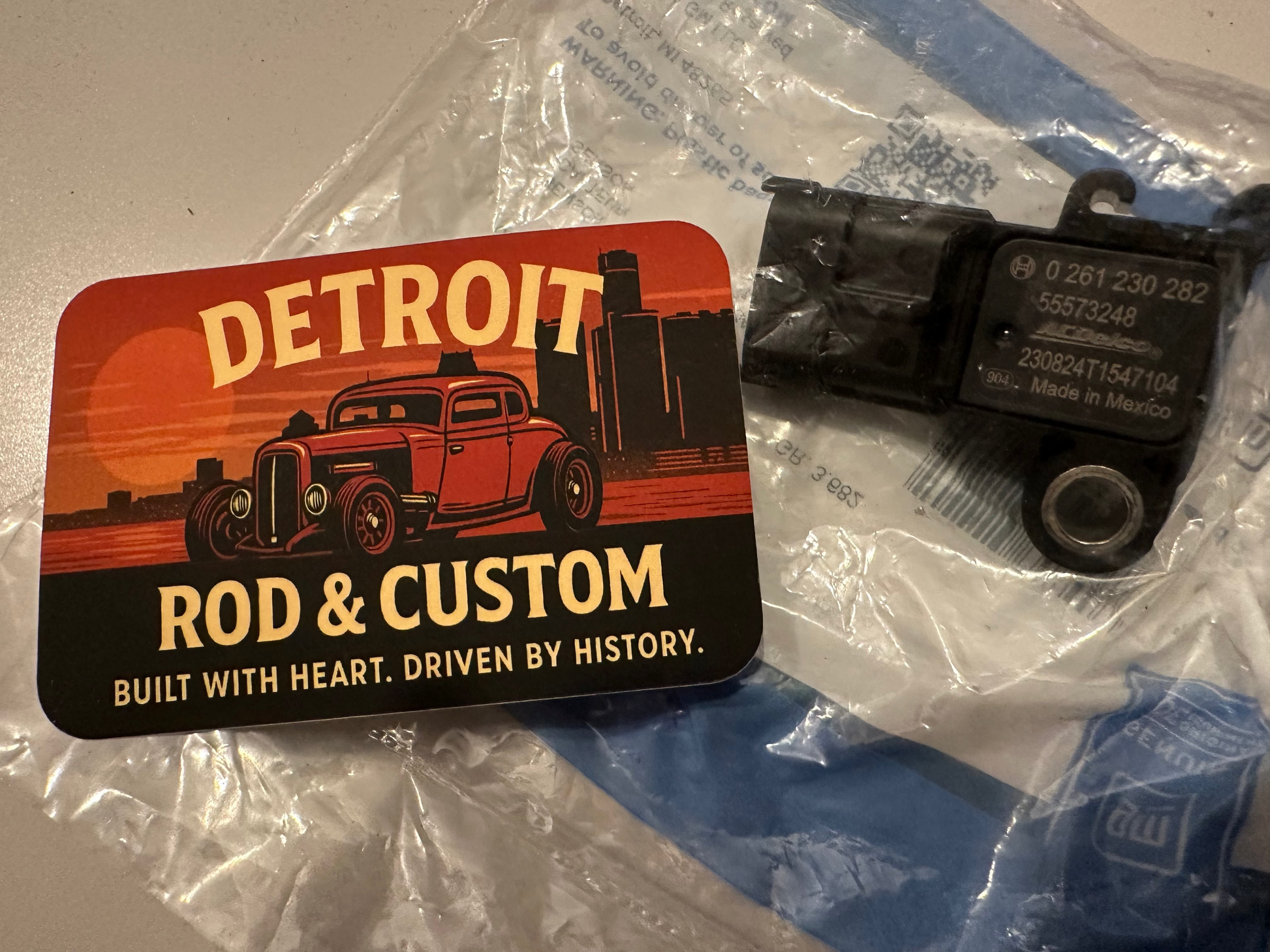 Mechanical component on a clear plastic sheet with 'Rod & Custom Detroit' branding.