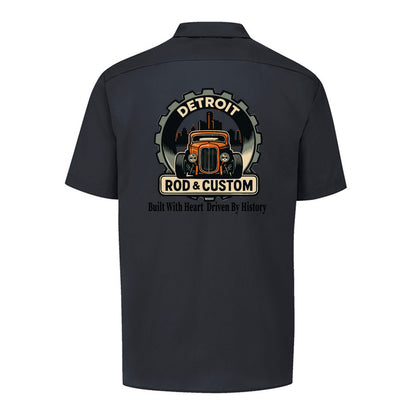 Back of a black Dickies short-sleeve work shirt with an orange hot rod coupe in a DETROIT ROD & CUSTOM gear badge over the Detroit skyline, with Built With Heart Driven By History below.