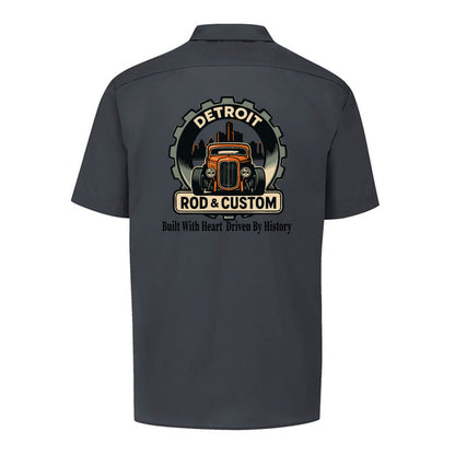 Back of a dark charcoal Dickies short-sleeve work shirt with an orange hot rod coupe in a DETROIT ROD & CUSTOM gear badge over the Detroit skyline, with Built With Heart Driven By History below.