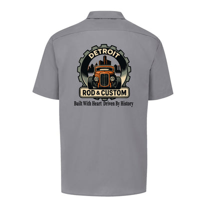 Back of a graphite grey Dickies short-sleeve work shirt with an orange hot rod coupe in a DETROIT ROD & CUSTOM gear badge over the Detroit skyline, with Built With Heart Driven By History below.
